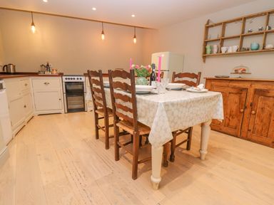 A kitchen with a dining table set with plates and candles wooden chairs white cupboards a wooden cabinet a fridge and hanging shelves at Bridge Studio in Broad Chalke