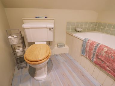 A bathroom with a toilet that has a wooden seat next to a bathtub with a tiled edge and a towel hanging at Bridge Studio in Broad Chalke
