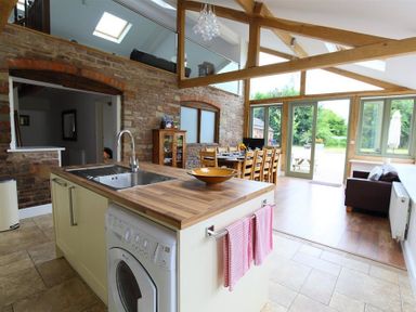 A kitchen island with a sink and washing machine next to a dining table and chairs in a room with exposed brick walls and wooden beams at The Old Mill in Flaxley