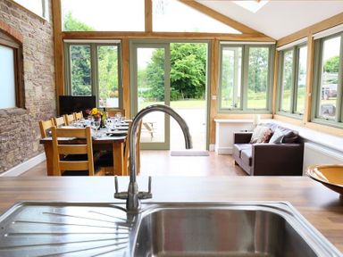 A kitchen sink with a dining table and chairs next to a stone wall and a sofa by windows at The Old Mill in Flaxley