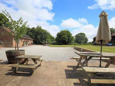 An outdoor patio area with wooden picnic tables one with a closed umbrella a gravel courtyard and a brick building surrounded by trees at The Old Mill in Flaxley