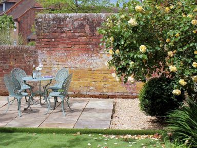 A patio with a metal table and four chairs with a bottle and glass on the table next to a brick wall and a blooming rose bush at Pops Barn in Downton