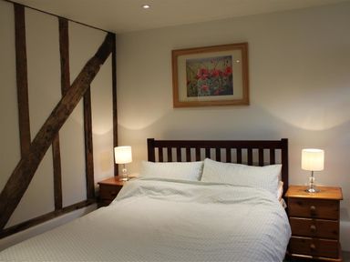 A bedroom with a double bed flanked by two wooden bedside tables with lamps and a framed flower picture on the wall at Pops Barn in Downton