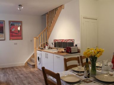 A dining table set with plates glasses and a vase of yellow flowers next to a kitchen area with white cabinets a washing machine and wooden staircase at Rojoy in Salisbury