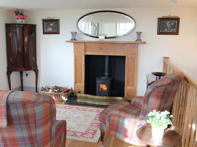 A sitting area with plaid armchairs, a fireplace with firewood basket, an oval mirror, paintings, and a cabinet at The Folly at Castlebridge in Mere
