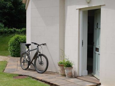 A bicycle leaning against a light-colored wall near a partially open door with two potted plants on a stone pathway at West Lodge in Rockbourne