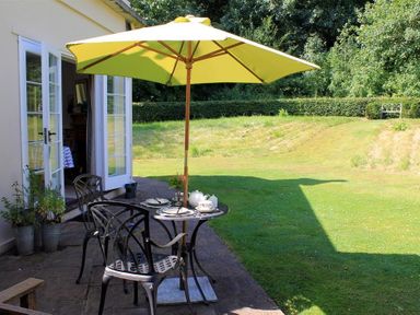 A patio with a table set for tea under a yellow umbrella and two metal chairs outside a house with open French doors at West Lodge in Rockbourne