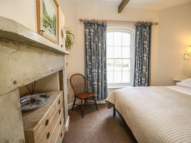 A bedroom with a bed covered in a beige quilt near a window with blue floral curtains and a wooden chair next to a white dresser at West Lodge in Rockbourne