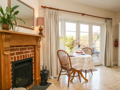 A dining area with a table covered with a checkered tablecloth and two chairs near glass doors and a fireplace with a wooden mantel at West Lodge in Rockbourne