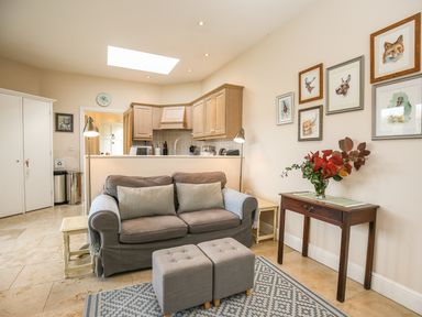 A sitting area with a sofa, two ottomans, a wooden console table with flowers, and a kitchen in the background at West Lodge in Rockbourne
