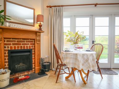 A dining area with a round table covered by a checkered tablecloth with two chairs near a fireplace and glass patio doors at West Lodge Rockbourne