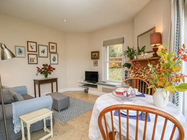 A living room with a sofa table with flowers and animal pictures on the wall a dining table with a vase of leaves and plates a television and plants on a wooden mantel at West Lodge Rockbourne
