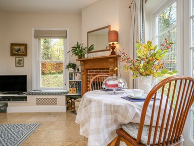 A dining table with a vase of leaves and a cup near a window and a fireplace with a mirror and lamp in a living room at West Lodge in Rockbourne