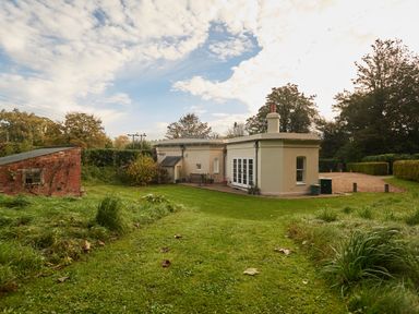 A garden with a green lawn and shrubs next to a small house and a red brick shed at West Lodge in Rockbourne