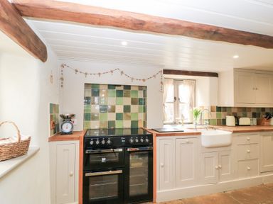 A kitchen with a black stove and oven, a white farmhouse sink, wooden countertops, cream cabinets, and green tiled backsplash at Cosy Cot in Stapleford