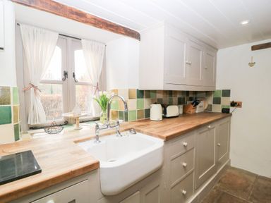 A kitchen with a white sink wooden countertops beige cabinets green and beige tiled backsplash and a window with white curtains at Cosy Cot in Stapleford