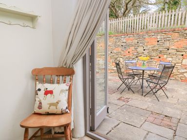 A wooden chair with a dog print cushion inside next to an open door leading to a stone patio with a table and four chairs at Cosy Cot in Stapleford