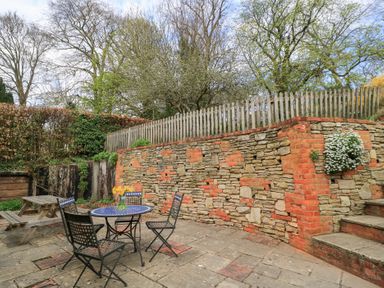An outdoor patio area with a stone and brick wall wooden picnic bench a round table with four chairs and flowers in a vase at Cosy Cot in Stapleford