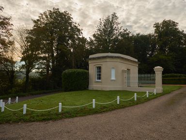 A small beige building with a window and gate on a grassy area near a gravel path and trees at East Lodge in Rockbourne
