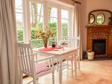 A dining area with a white wooden table and chairs with red and white checkered cushions by large windows and a fireplace with a wooden mantle at East Lodge in Rockbourne