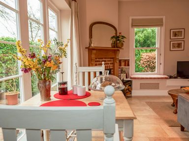 A dining area with a wooden table and white chairs next to windows with flowers and a cake cover at East Lodge in Rockbourne