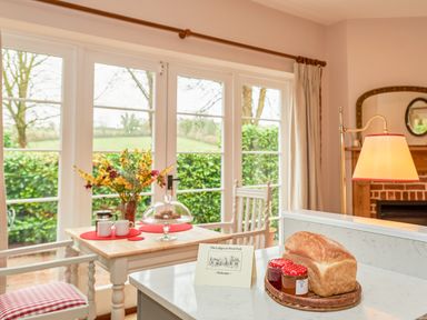 A kitchen counter with a loaf of bread and jam jars next to a welcome card and a dining table with cups and flowers at East Lodge in Rockbourne