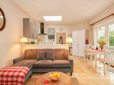 A living room with a gray sofa a round wooden table a kitchen area and a dining table with flowers at East Lodge Rockbourne
