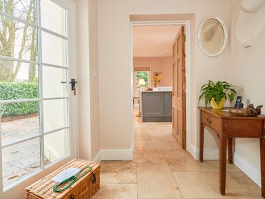 A hallway with a wicker basket on the floor a wooden table with a plant a phone and a decorative item a round mirror on the wall and a view into a kitchen at East Lodge in Rockbourne