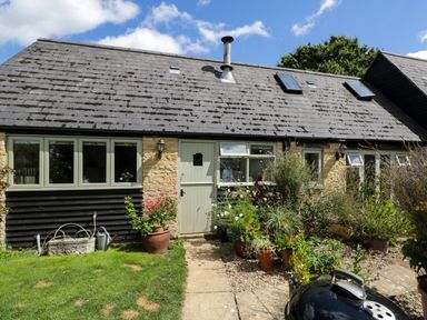 Exterior of a stone and wood house with a front door windows potted plants and a charcoal grill at Henmarsh Cottage in Moreton-In-Marsh