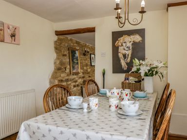 A dining room with a set table featuring bowls cups and a teapot with dog and rooster artwork on the walls at Henmarsh Cottage in Moreton-In-Marsh