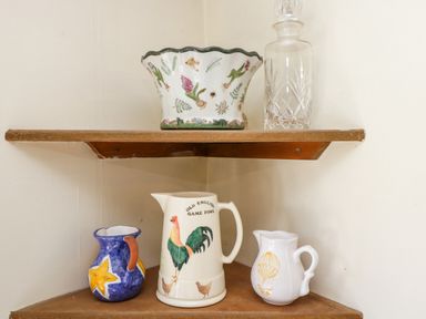 A corner shelf with a decorative bowl and glass bottle on the top shelf and three painted ceramic pitchers on the bottom shelf at Henmarsh Cottage in Moreton-In-Marsh