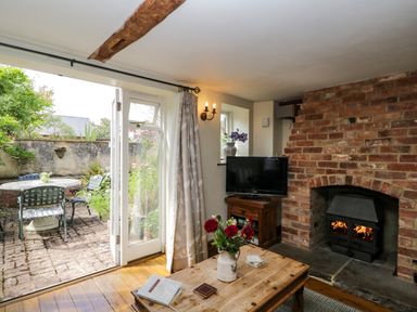 A living room with a brick fireplace and wooden table next to an open door leading to a garden at Henmarsh Cottage in Moreton-In-Marsh