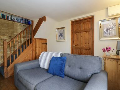 A living room with a gray sofa a blue pillow wooden stairs and wooden door at Henmarsh Cottage in Moreton-In-Marsh