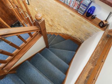 A staircase with blue carpet and wooden railing next to a stone wall with a wooden shelf holding books and decorative items at Henmarsh Cottage in Moreton-In-Marsh