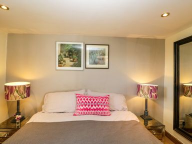 A bedroom with a bed a pink patterned pillow two table lamps and two framed pictures above the bed at Henmarsh Cottage in Moreton-In-Marsh