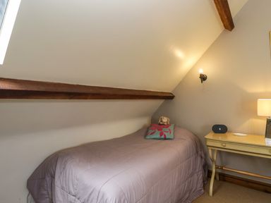 A bedroom with a single bed covered with a lavender quilt a pillow with a stuffed toy a yellow bedside table with a lamp and a speaker at Henmarsh Cottage in Moreton-In-Marsh