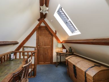 An attic room with a wooden door wooden beams a desk with chair a large wooden chest a small side table lamp and skylight at Henmarsh Cottage in Moreton-In-Marsh