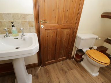 A bathroom with a white sink and soap dispenser next to a wooden door and a toilet with a wooden seat at Henmarsh Cottage in Moreton-In-Marsh