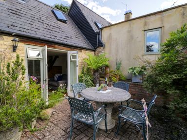 An outdoor patio area with a round stone table and four metal chairs surrounded by potted plants and a stone building with open double doors at Henmarsh Cottage in Moreton-In-Marsh