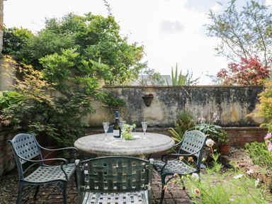 An outdoor patio with a round stone table and four metal chairs surrounded by plants and trees at Henmarsh Cottage in Moreton-In-Marsh