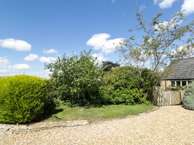 A garden with bushes trees a gravel driveway and a wooden gate at Henmarsh Cottage in Moreton-In-Marsh