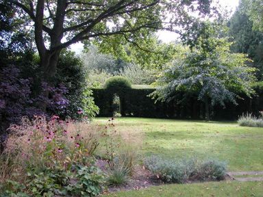 A garden with trees shrubs and a hedge with an arch opening at The Yeoman's House in Bignor