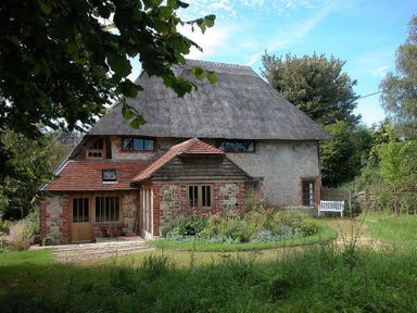 A thatched roof house with brick and stone walls surrounded by trees and a garden with a white bench at The Yeoman's House in Bignor