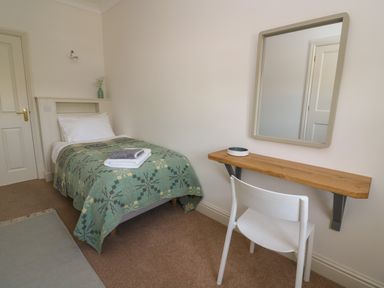 A single bed with patterned blanket and towels on top next to a wooden wall-mounted desk with a white chair and a square mirror in a bedroom at Craigluscar in Newport Pembrokeshire