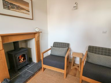 A room with two wooden armchairs with cushions a small wooden stool a wood burning stove in a wooden fireplace and a framed picture on the wall at Craigluscar in Newport Pembrokeshire