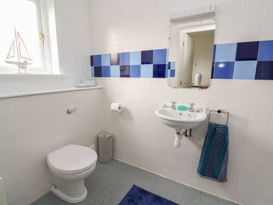 A bathroom with a toilet, wall-mounted sink, mirror, towel rack with cloth, wastebasket, and window with a small sailboat model at Craigluscar in Newport, Pembrokeshire