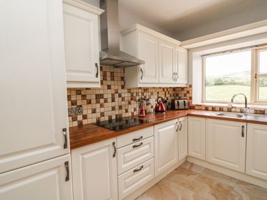 A kitchen with white cabinets brown tiled backsplash wooden countertops a stovetop a stainless steel range hood a red kettle a toaster a window over the sink showing a grassy landscape at Dromstabla in Glencar