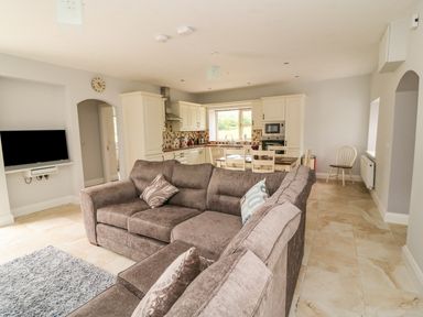 An open plan living area with a brown sectional sofa a wall mounted TV a dining table and chairs and a kitchen with white cabinets at Dromstabla in Glencar