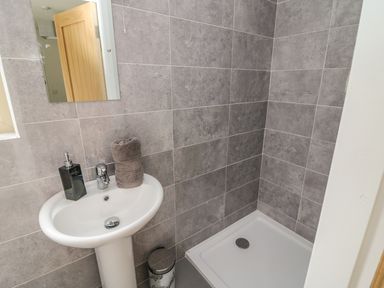 A bathroom with a pedestal sink a mirror a towel liquid soap dispenser a trash bin and a shower tray with gray tiled walls at Dromstabla in Glencar