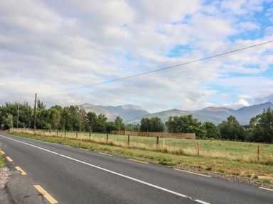 A paved road beside a fenced grassy field with trees and mountains in the background at Dromstabla in Glencar